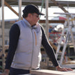 Person wearing a white ac wear fan cooled jacket air conditioned vest with a logo, standing outdoors near construction materials.