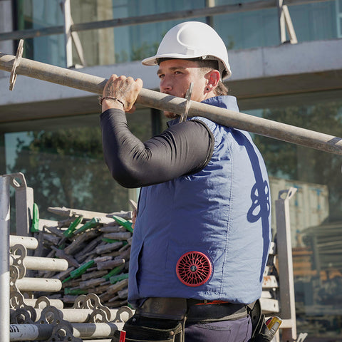 Person wearing a hard hat and purple fan cooled jacket carrying a long metal rod on a construction site.